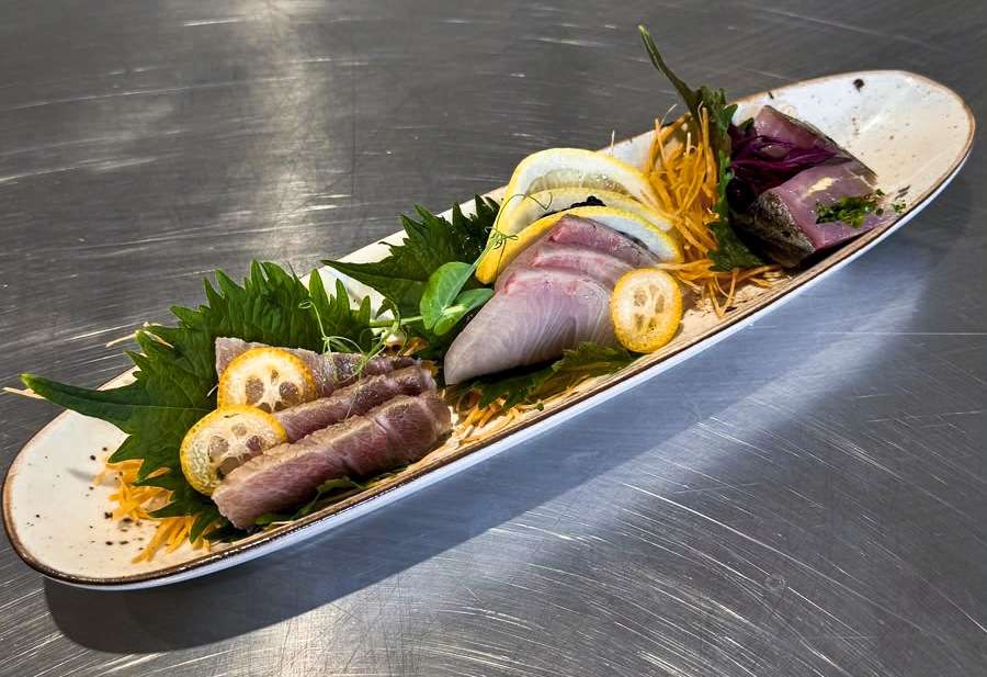 Long platter of o-toro, akami and kinmedai sashimi with citrus and leaves on a steel counter