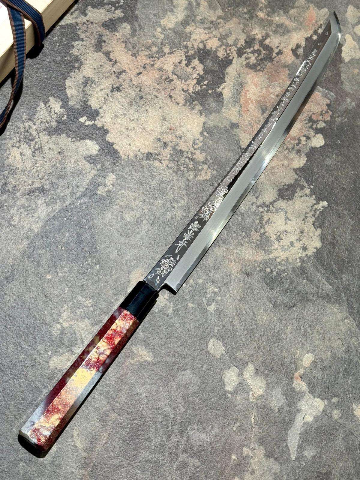Long-bladed yanagiba knife with a lacquered handle resting diagonally on a stone surface